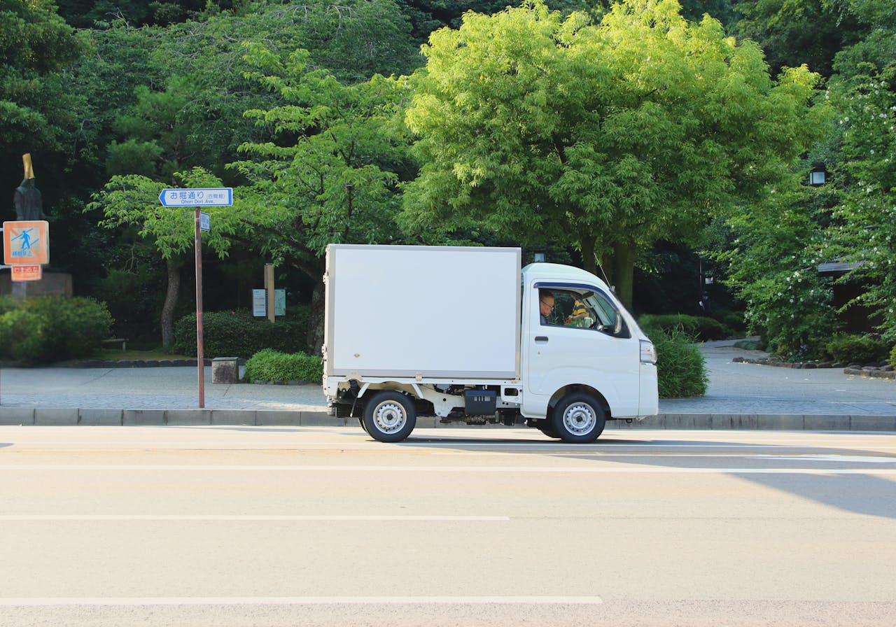 White delivery truck driving on a sunny day in Hiroshima, Japan, surrounded by green trees.